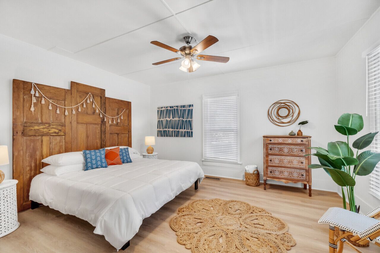 A bedroom with a wooden headboard, a ceiling fan, a bed with white bedding, a woven rug, a wooden dresser, and a large green plant.