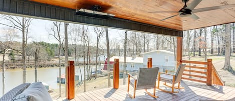 Um deck de madeira com móveis de exterior, um ventilador de teto e vista para um lago e edifícios ao fundo.