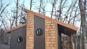 A small wooden cabin with a metal roof, two circular windows, and a wooden staircase leading to a porch.