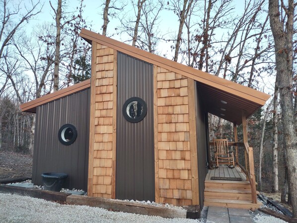 A small wooden cabin with a metal roof, two circular windows, and a wooden staircase leading to a porch. - Owl house was built for unplugging, relaxing & connecting with friends/family (Troy)