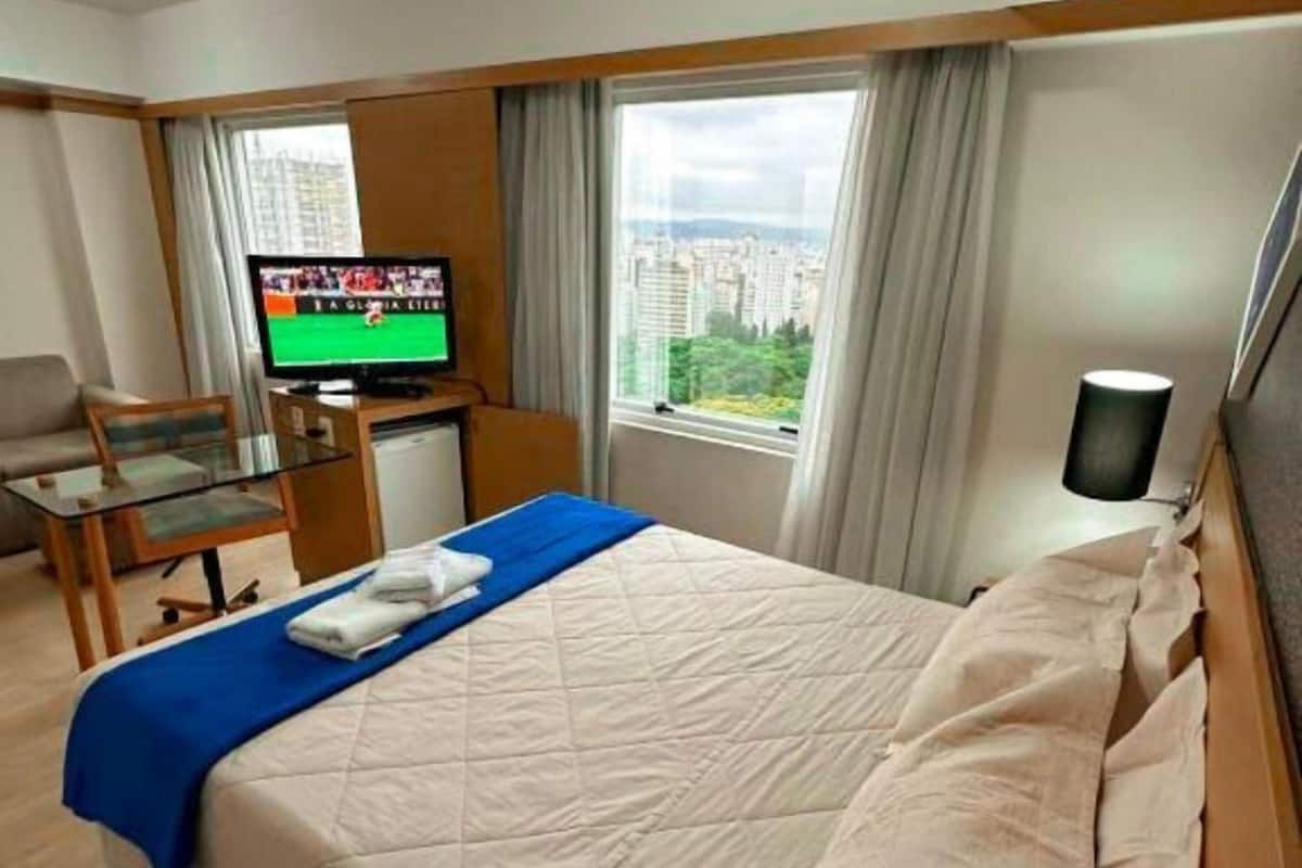 A hotel room with a bed, a television, a desk, and a view of the city.