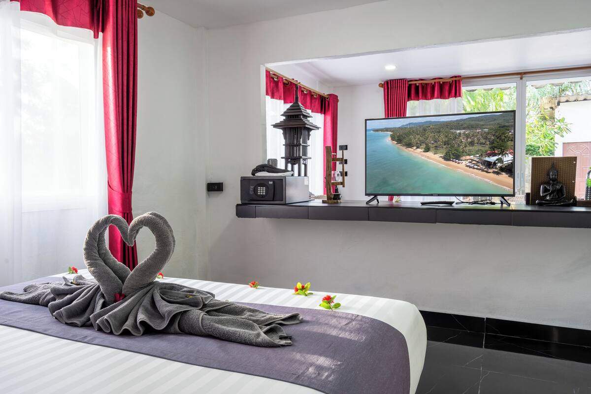 Habitación de hotel con cama, televisor mostrando una vista de playa, un pequeño santuario y una cortina roja y blanca.