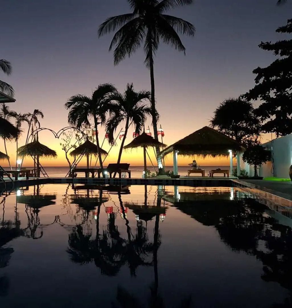 A resort pool area with palm trees and thatched umbrellas, reflecting the sunset.