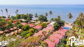 Un resort con varias edificaciones, algunas con techos rojos, rodeado de exuberante vegetación y palmeras, con vista a un mar tranquilo.