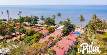 Un resort con varias edificaciones, algunas con techos rojos, rodeado de exuberante vegetación y palmeras, con vista a un mar tranquilo.