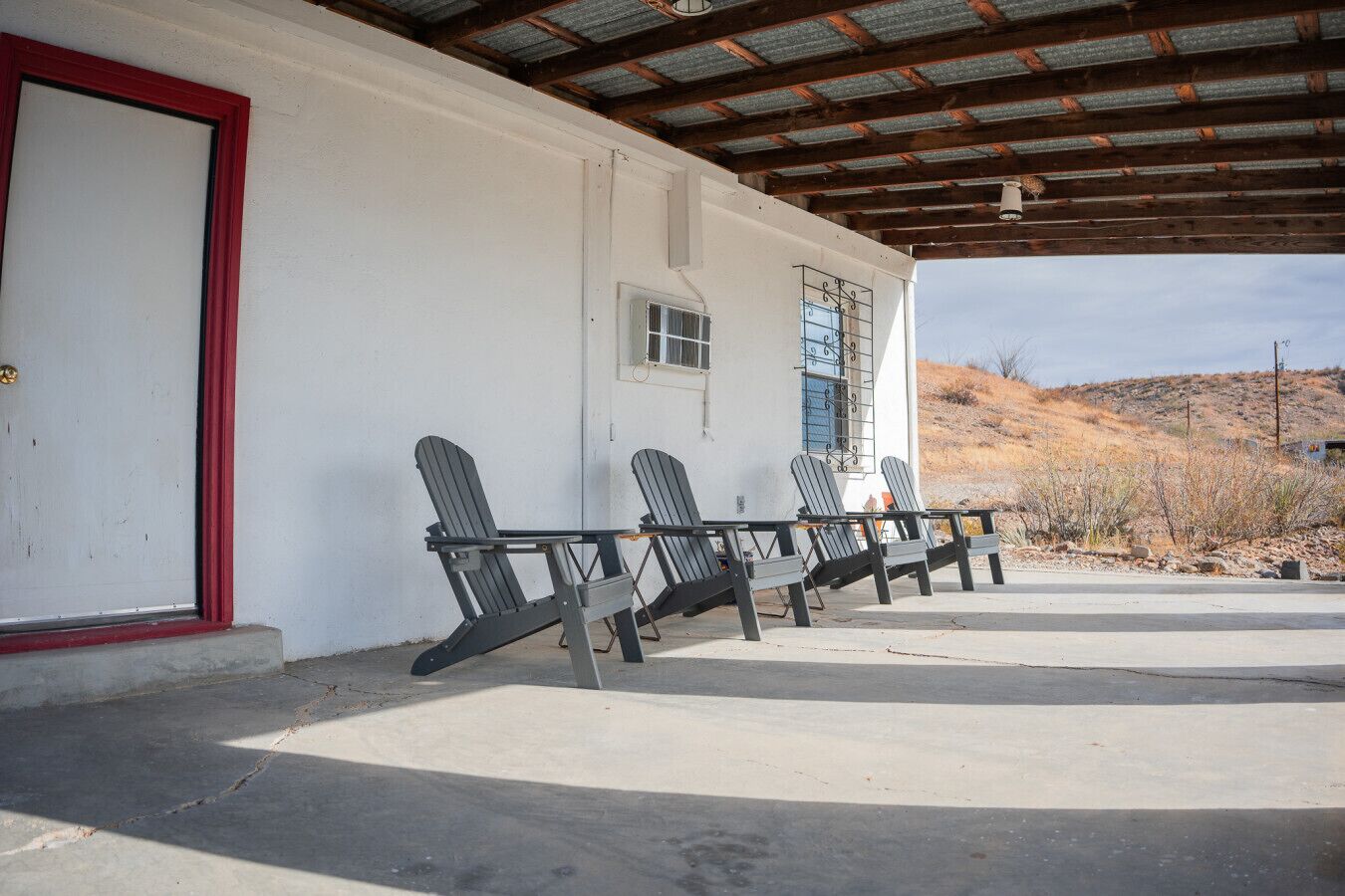 Un espace extérieur abrité avec des chaises Adirondack, un bâtiment blanc avec une porte rouge et un toit en métal.