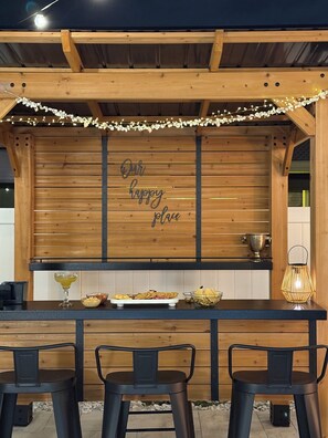 Un área de bar exterior con barra de madera, un letrero que dice 'Our Happy Place', taburetes de bar, una copa de bebida, aperitivos y un farol decorativo.