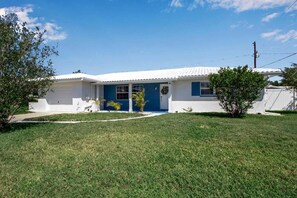 Una casa de un solo piso con fachada blanca y detalles azules, una puerta principal y un pequeño jardín delantero con césped y algunas plantas.