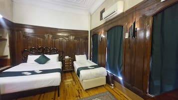 Quarto de hotel com duas camas, um guarda-roupa de madeira, uma pequena mesa de cabeceira e um tapete estampado.