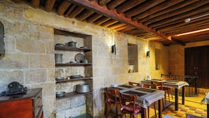 Un comedor rústico con paredes de piedra, vigas de madera y suelo a cuadros. Las mesas están dispuestas con sillas, y hay estantes con ollas y sartenes de metal.