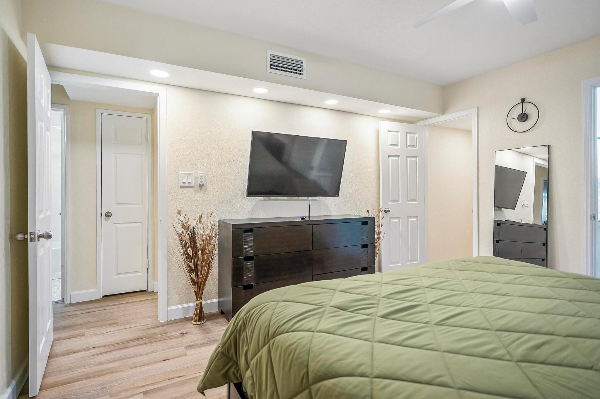 A bedroom with a bed, a TV mounted on the wall, a dresser, and a door leading to another room.
