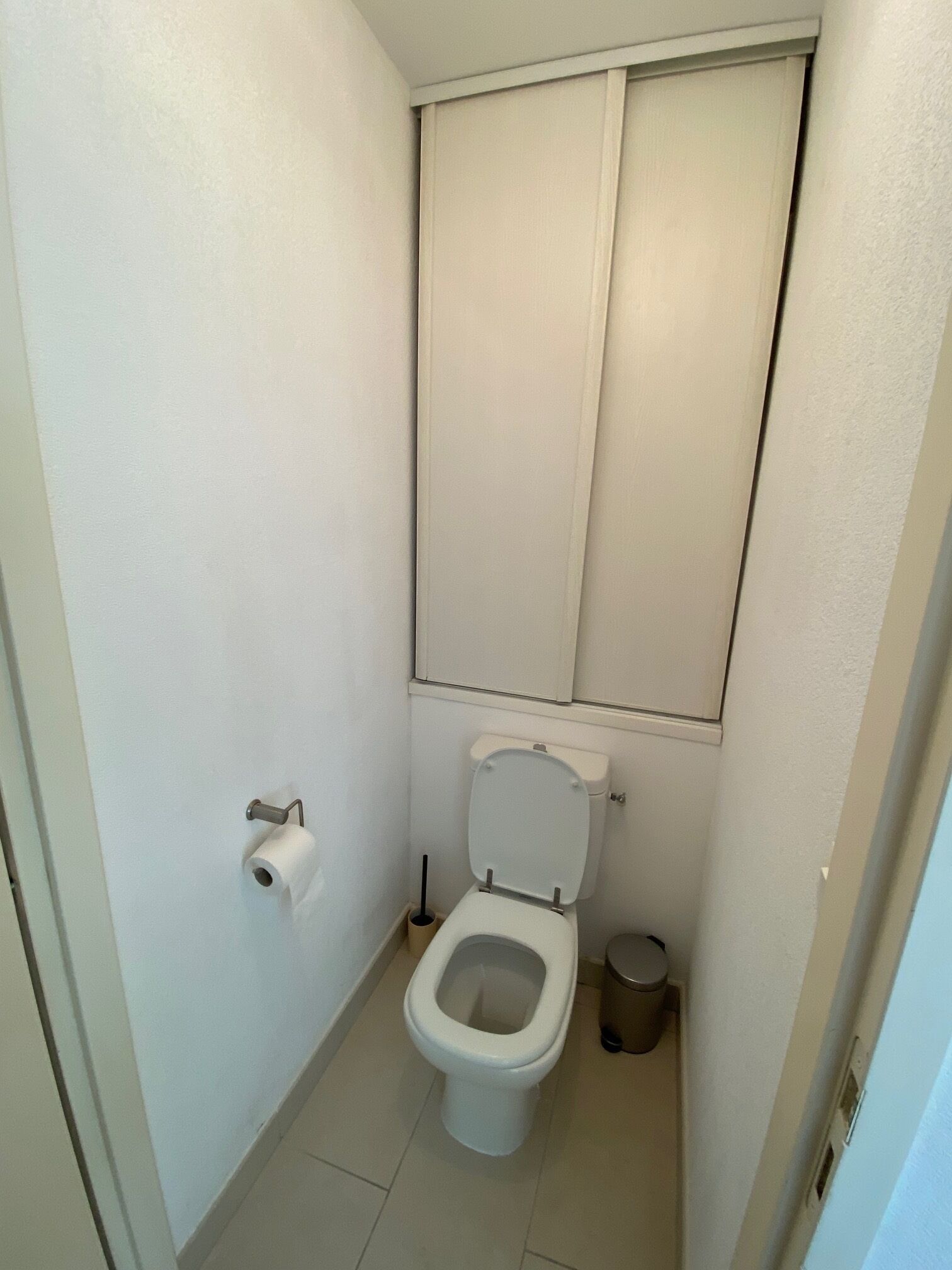 Un baño pequeño y funcional con inodoro, dispensador de papel higiénico, cubo de basura y ventana con persianas.