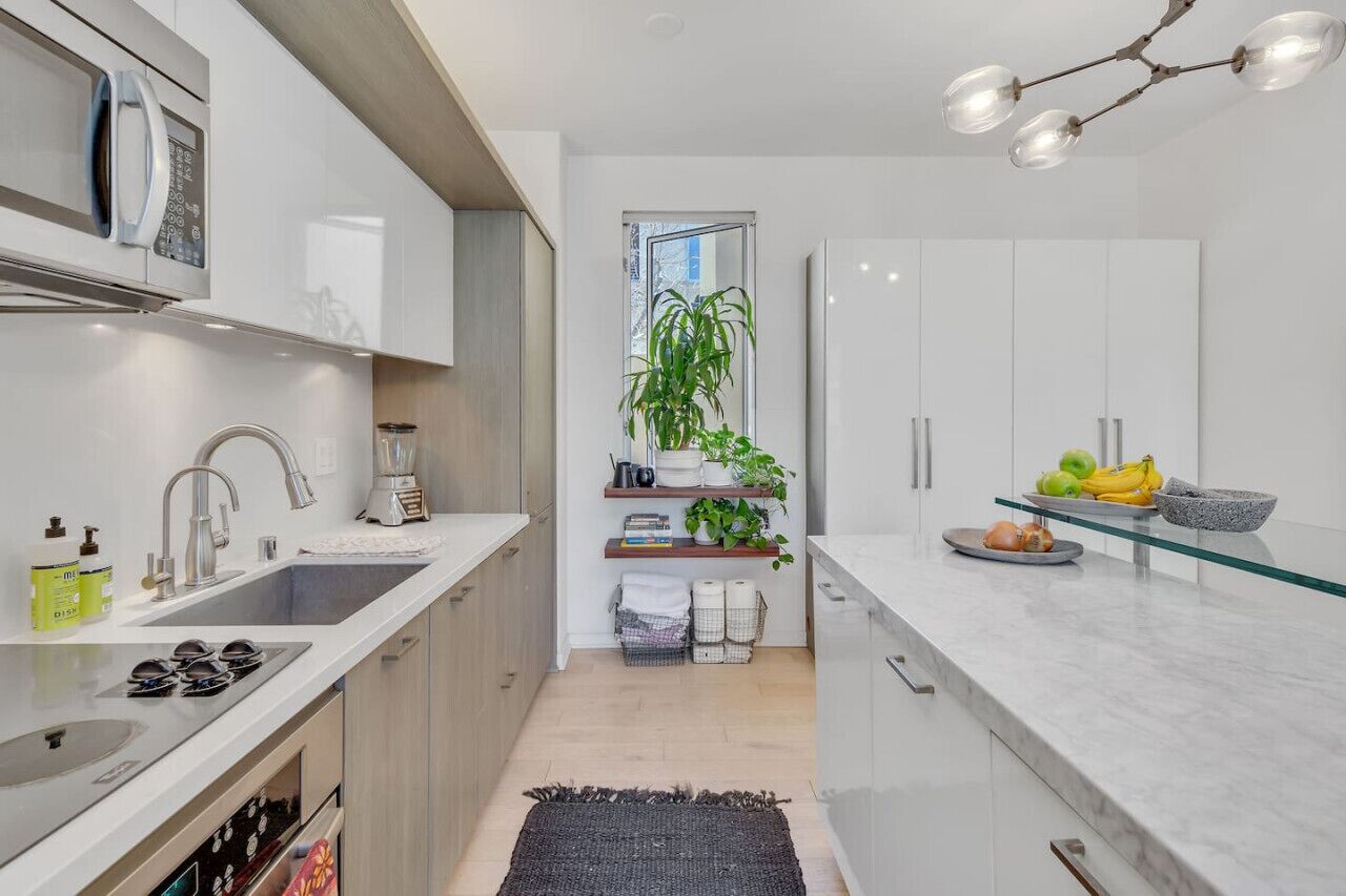 A modern kitchen with a built-in microwave, stainless steel sink, and a glass countertop. There is a plant shelf and a fruit bowl on the counter.