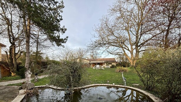 A circular pond with a wooden deck edge, surrounded by trees and a grassy area with outdoor furniture.