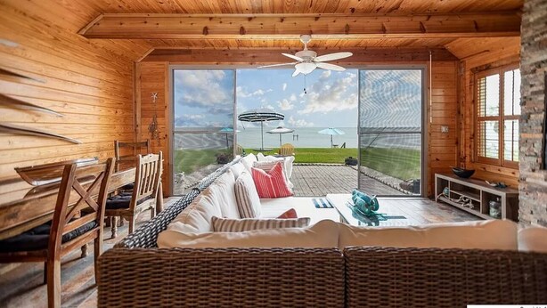 A wooden cabin interior with a large sliding glass door leading to an outdoor deck. Inside, there is a sofa, a dining table, and a ceiling fan. - We Dig It at Magnolia Beach Texas (Port Lavaca)