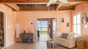 A spacious living room with a wooden ceiling, a white sofa, a striped ottoman, and a view through an open door to a balcony. - We Dig It at Magnolia Beach Texas (Port Lavaca)