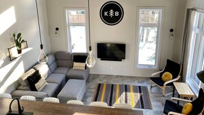 A modern living room with a grey sectional sofa, a flat-screen TV mounted on the wall, and a dining area with a wooden table and chairs.