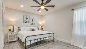 Un dormitorio con cama, mesitas de noche, ventilador de techo y cuadros en la pared.
