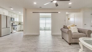 Un salón amplio con un sofá gris, una mesa de centro de vidrio y un ventilador de techo. Se ve una cocina con armarios y electrodomésticos blancos al fondo.