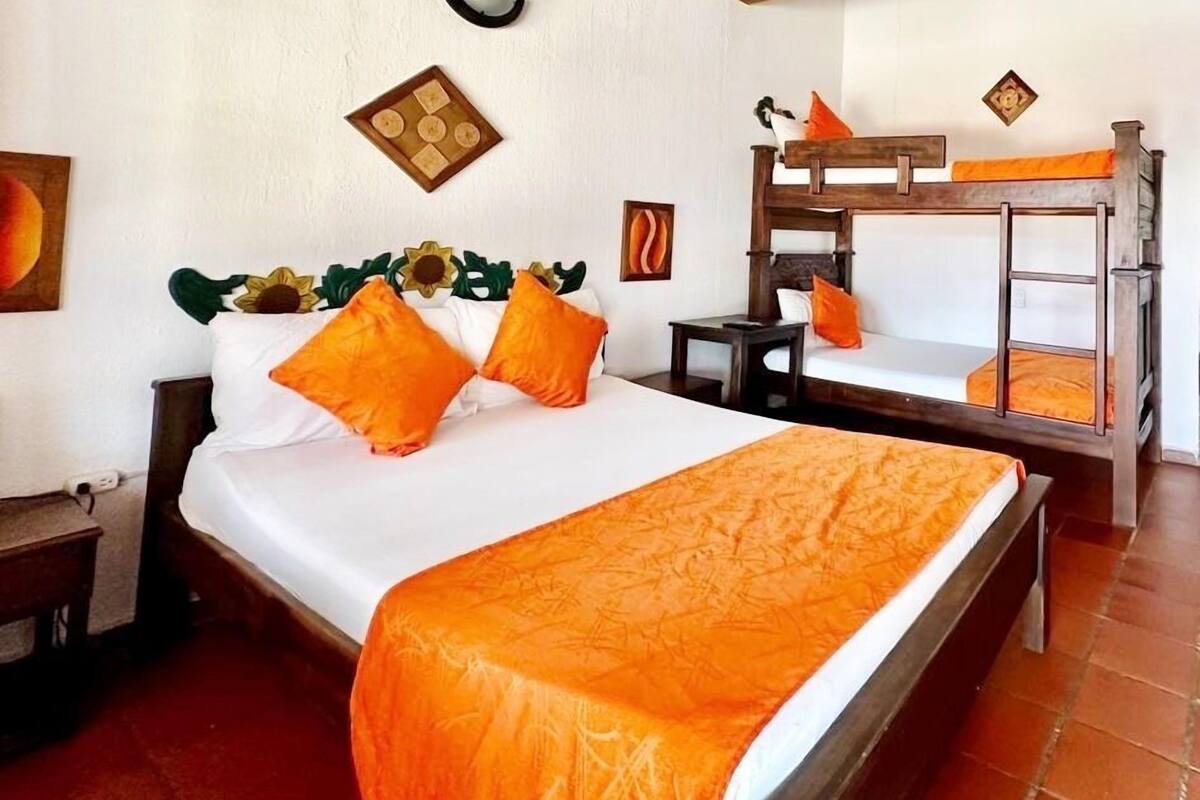 A room with a large bed, a smaller bed, a nightstand, a wooden bench, and a wall-mounted light fixture.