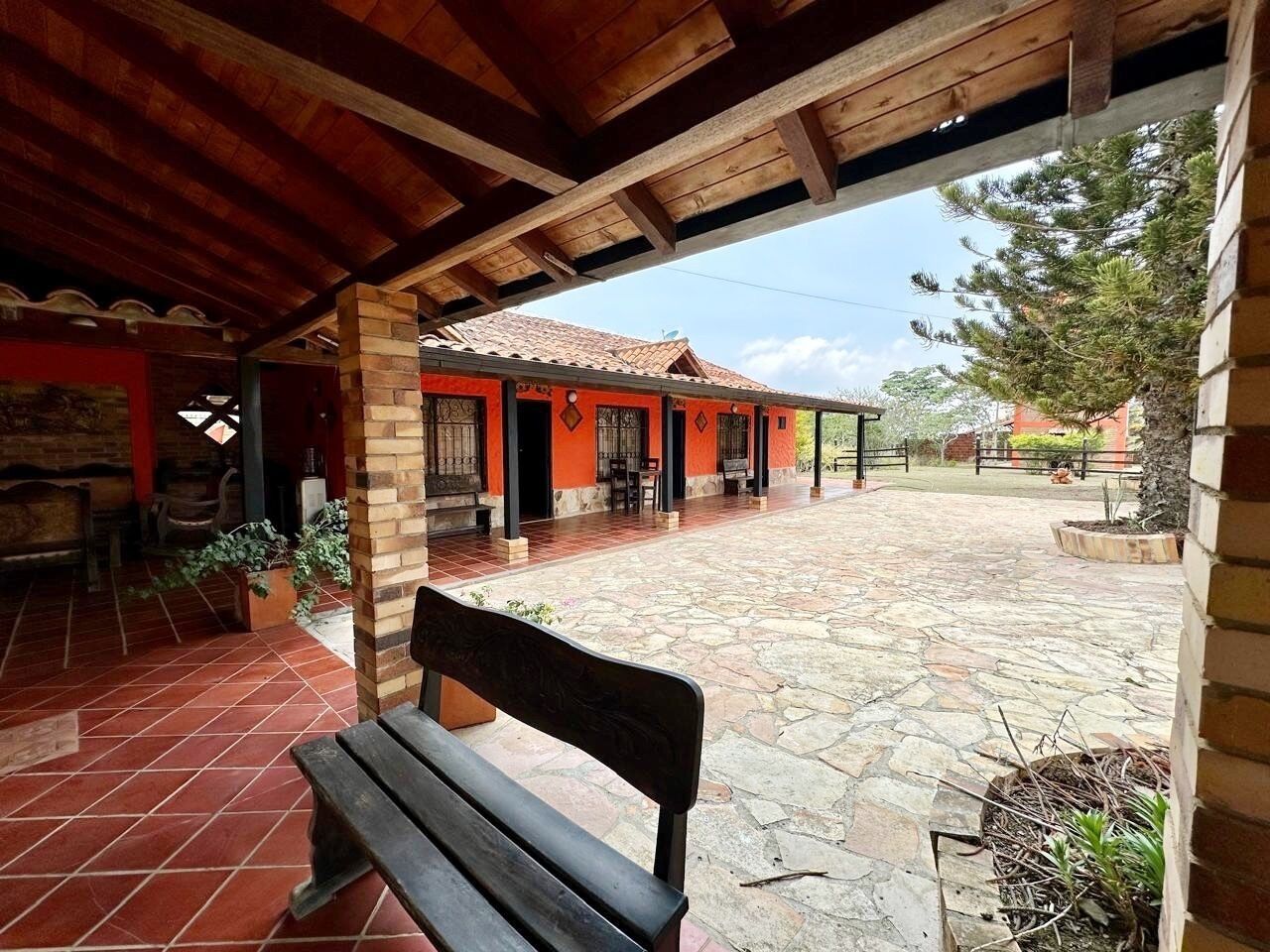 A covered patio area with a stone floor, a wooden bench, and a building with red walls and a tiled roof.