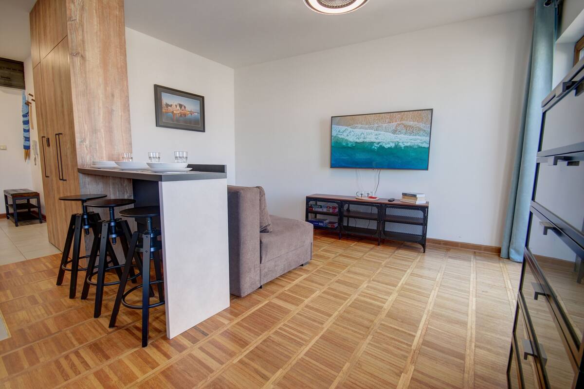 A modern kitchen with a central island, bar stools, a sofa, and a large TV mounted on the wall.