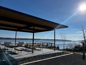 Un espace extérieur aménagé avec des tables et des bancs, un pavillon couvert et une vue sur un lac et des reliefs lointains.