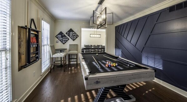 A pool table in a room with a dark accent wall, a bar area, and a seating area with a chair and a table.