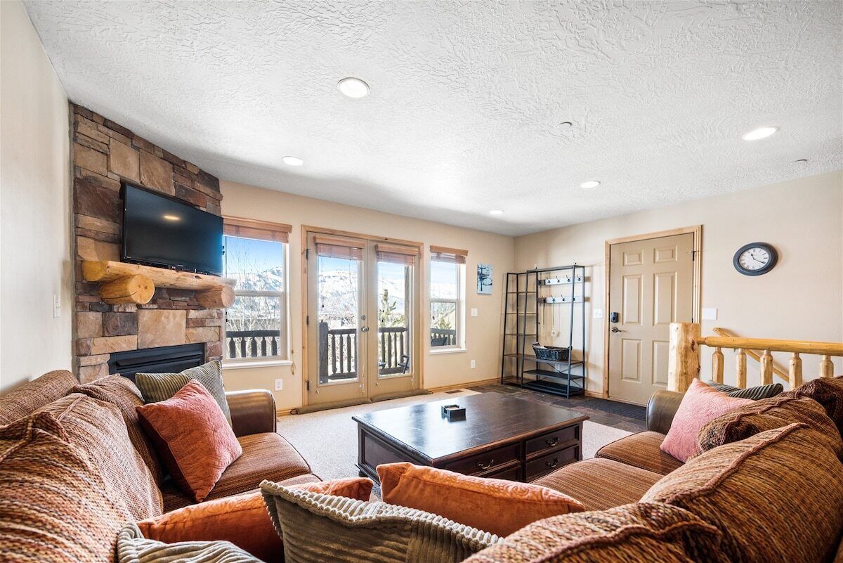 A cozy living room with a stone fireplace, a flat-screen TV, and a large window overlooking a deck.