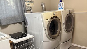 A laundry room with two front-loading washing machines, a drying rack, a clock on the wall, and a sign indicating the room is available for rent. - Canyon View (Lewiston)