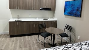 A compact kitchen with dark wooden cabinets, a white countertop, and a black range hood. There is a small round table with two chairs, and a large abstract painting on the wall. - Palace Inn Blue Jones Rd  (Houston)