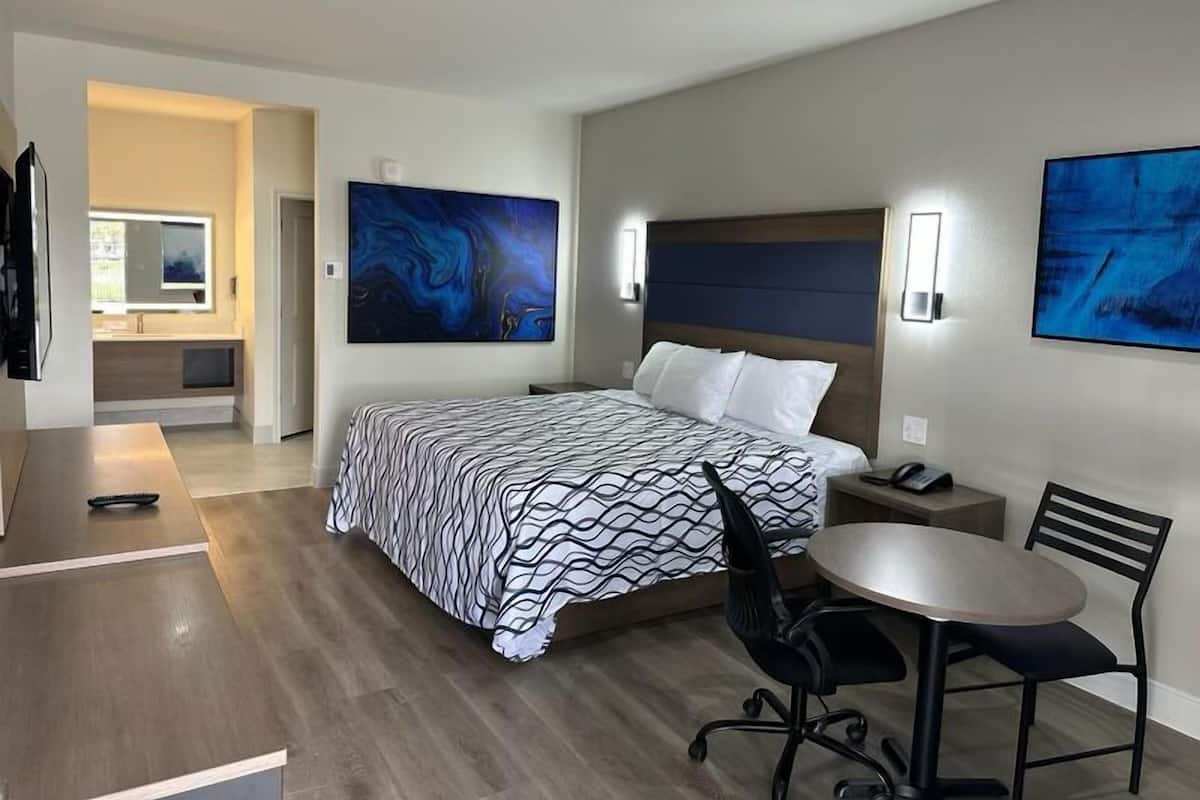 Et moderne hotelværelse med en stor seng, et skrivebord, en stol og et lille bord. Der er to billeder på væggene, og et badeværelse kan ses i baggrunden.