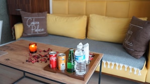Un coin salon accueillant avec un canapé jaune et une table basse en bois sur laquelle sont posés une bougie, des canettes et une bouteille d’eau.