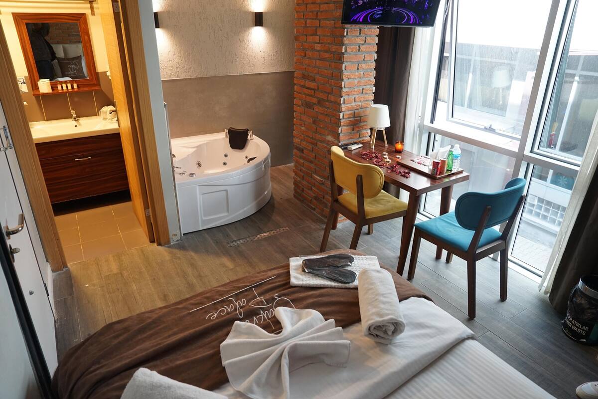A hotel room with a bed, a chair, a small table with toiletries, a TV, and a view of the outside.