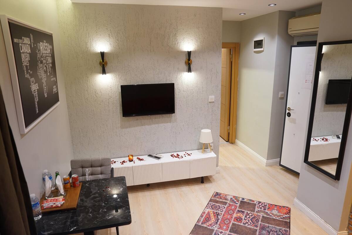 A hotel room with a flat-screen TV mounted on the wall, a white console table with a lamp, a checkered sofa, a small table with snacks and a bottle, and a patterned rug.