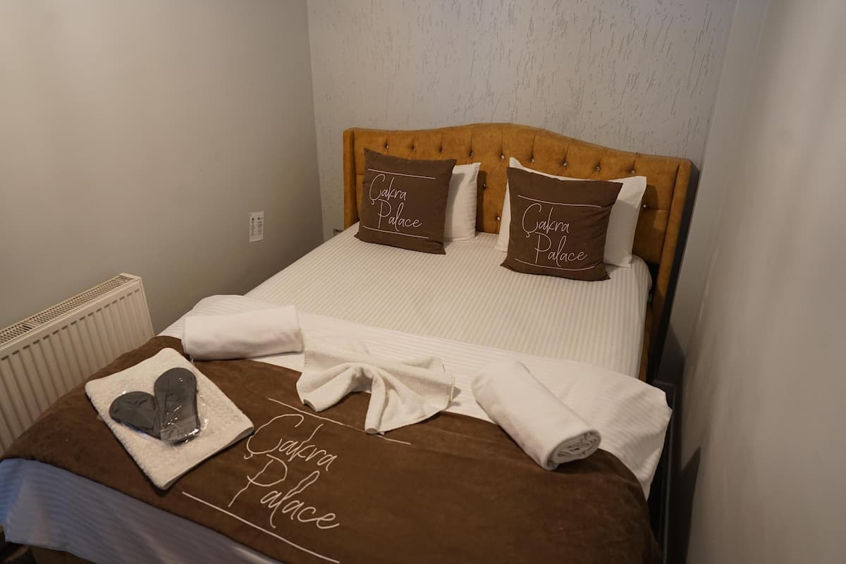 A neatly made bed with a brown headboard, two pillows with 'Gakra Palace' text, and a towel with a heart design.