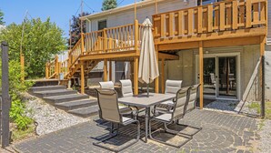 A patio with a table and chairs, a wooden deck, and a staircase leading to a balcony. - Green Lawn (Grawn)