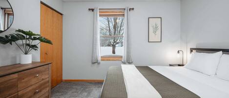 A bedroom with a bed, a dresser, a potted plant, a window with curtains, and a framed picture on the wall.