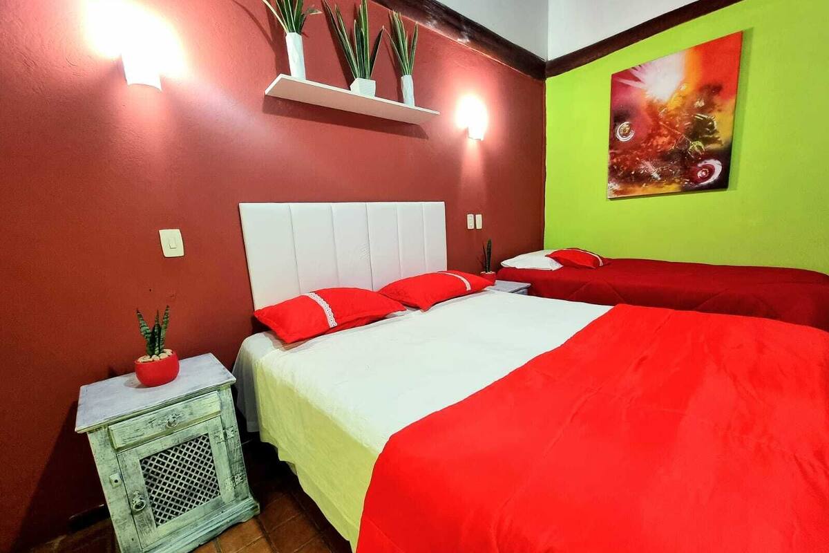 Una habitación con dos camas, ropa de cama roja, cabecera blanca, una mesita con una planta y una pared verde con un cuadro.