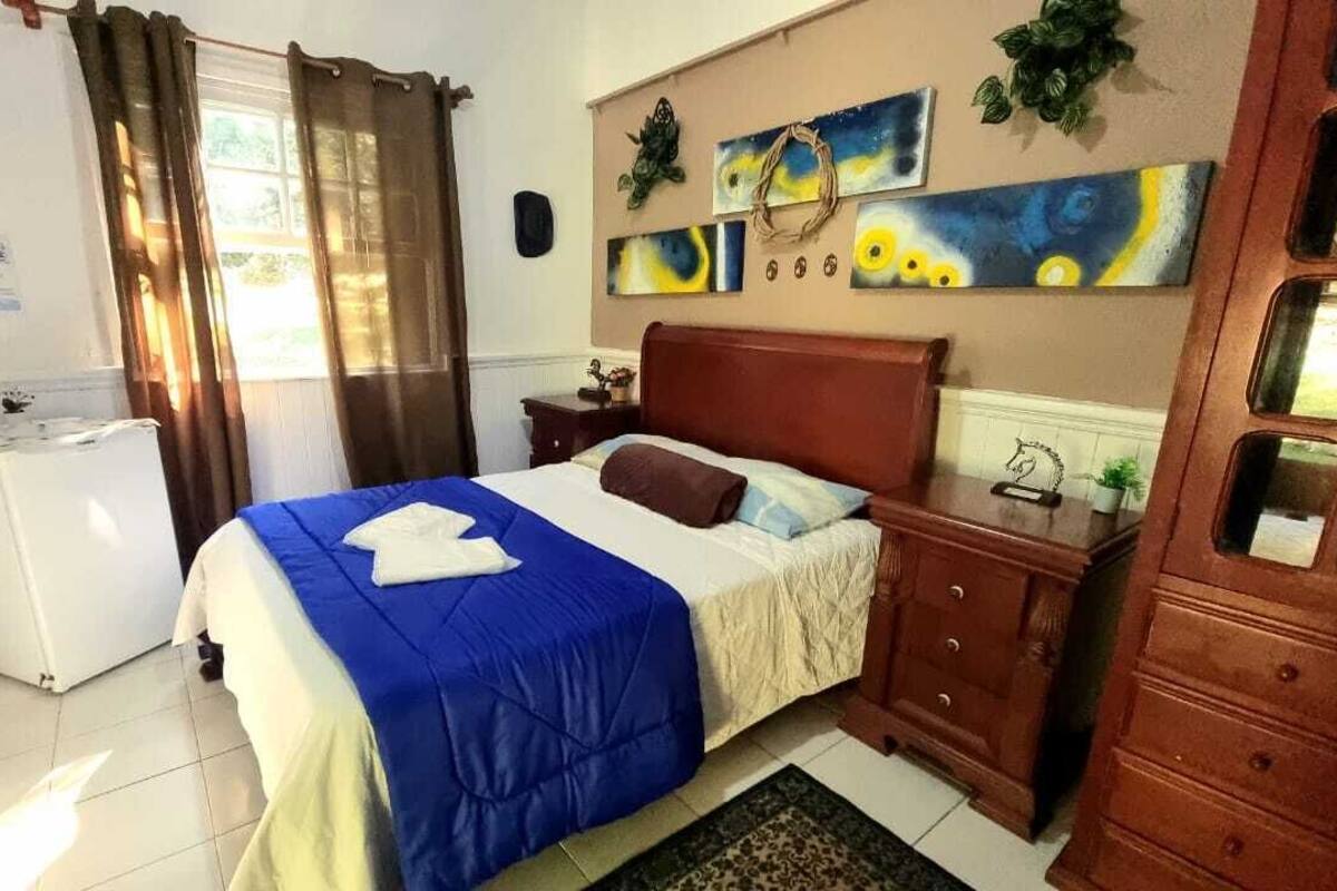 Un dormitorio con cama, mesita de noche, cómoda, refrigerador y ventana con cortinas.