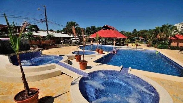 A pool area with multiple hot tubs and a small pool, surrounded by potted plants and a red-roofed structure. - Jaco Condo - Walk to beach! (Jaco)