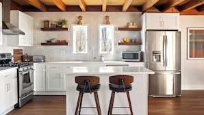 Une cuisine moderne avec des meubles blancs, des appareils en acier inoxydable, des poutres en bois au plafond et deux tabourets de bar.