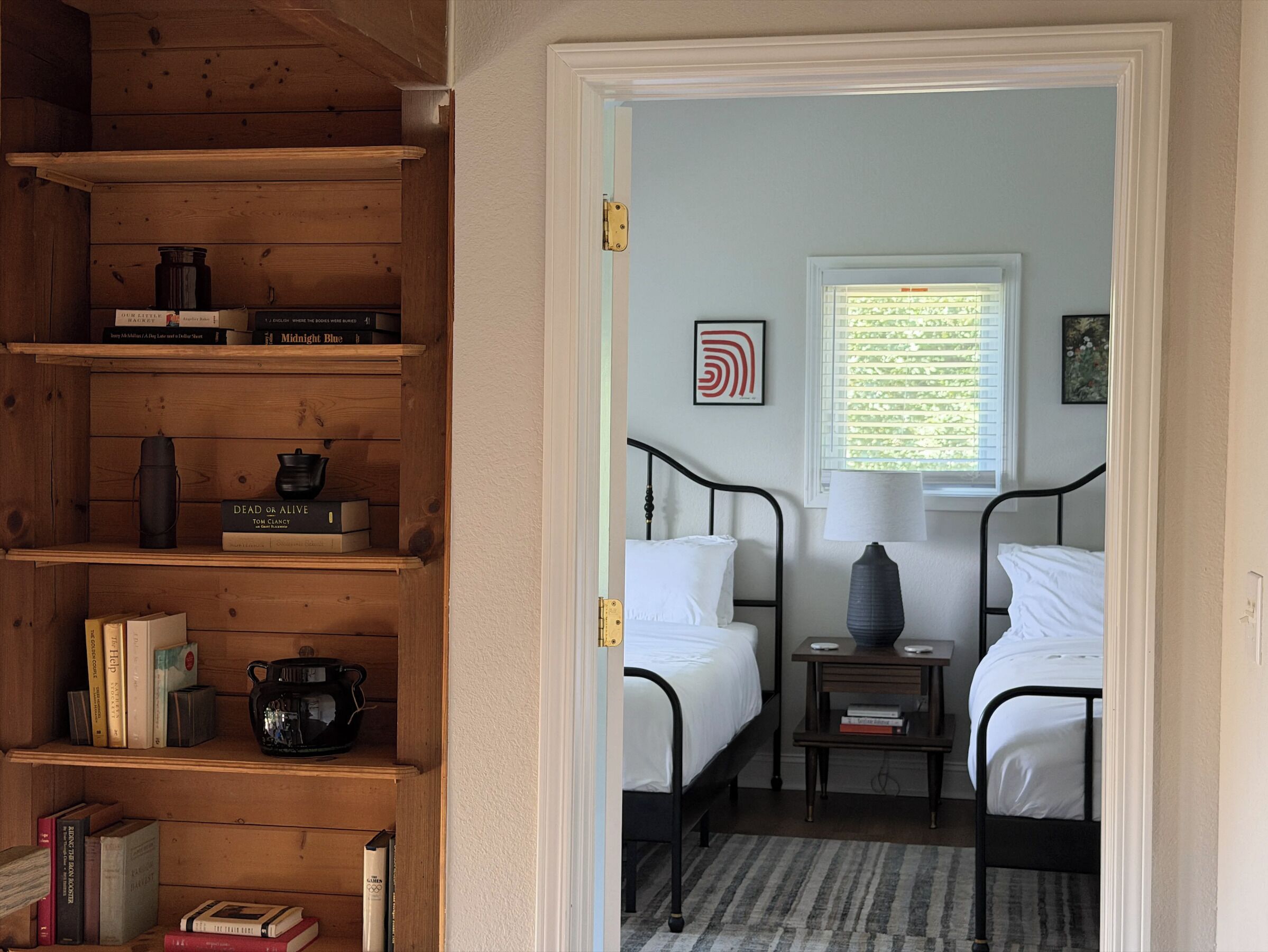 Une chambre avec deux lits, une table de chevet, une lampe, une fenêtre avec des stores, une bibliothèque et une porte.