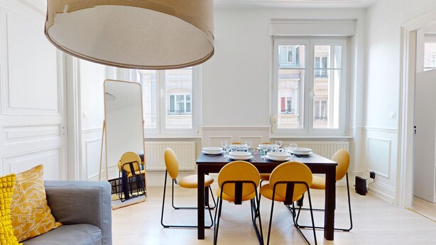 Un coin repas avec une table dressée pour quatre personnes, des chaises jaunes et un grand luminaire suspendu.