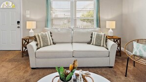Una sala de estar con un sofá color beige, dos mesitas de noche con lámparas, un sillón de mimbre y una ventana con cortinas.