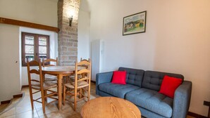 Una sala de estar con pared de piedra, una mesa de comedor de madera con sillas, un sofá azul con cojines rojos y una mesa de centro redonda de madera.