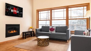 A cozy living room with a fireplace, a grey sofa, a coffee table, and a large window with a view of trees. - Serene, breathtaking waterfront views in Muskoka, half an hour from Algonquin! (Huntsville)