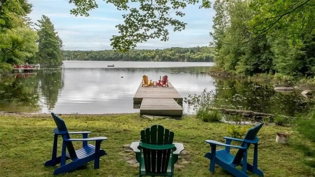 A lakeside deck with Adirondack chairs and a small table, overlooking a calm lake with a dock and boat in the distance. - Serene, breathtaking waterfront views in Muskoka, half an hour from Algonquin! (Huntsville)