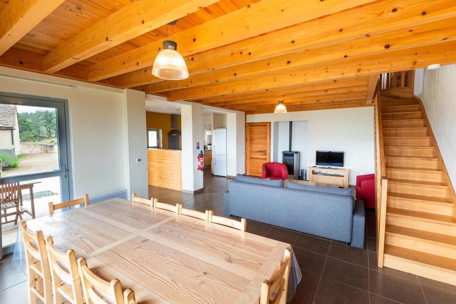 Un área de estar amplia con techo de madera, una mesa de comedor con sillas, una zona de cocina, un sofá gris, una butaca roja, un televisor y una escalera.