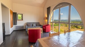 A spacious room with a large arched window, a grey sofa, a red armchair, and a wooden table. - La Baumelle 4/6 persons (SAINT-PIERRE-DES-TRIPIERS)